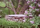 A stone well in front of a tree and behind some lilacs